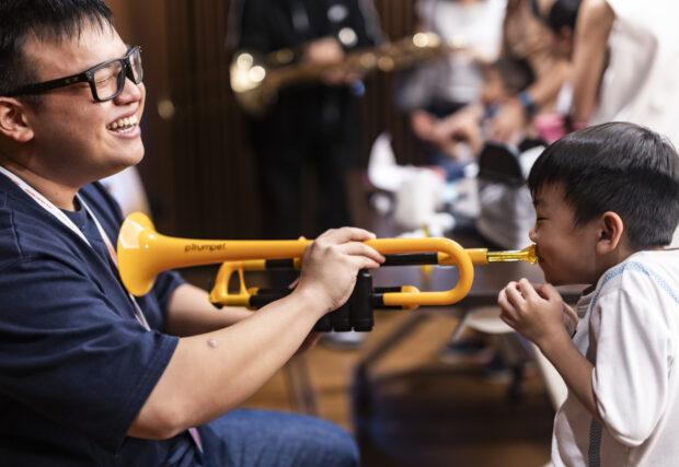 Trying a plastic trumpet at the Instrument Petting Zoo with the help of a student volunteer